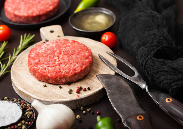 Fresh raw minced homemade farmers grill beef burger on round chopping board with spices and herbs and fork and knife on brown board. Oil, pepper and garlic. - Foto, imagen