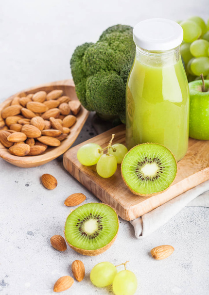 Glass bottle of fresh smoothie juice organic green toned fruit and vegetables on stone kitchen background. With almond nuts in bowl. - Fotoğraf, Görsel