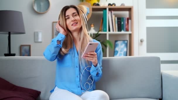 Portrait of young cute attractive young girl in cozy living room background listening to music with headphones. Woman wearing blue blouse enjoying the music and dancing at home. - Záběry, video