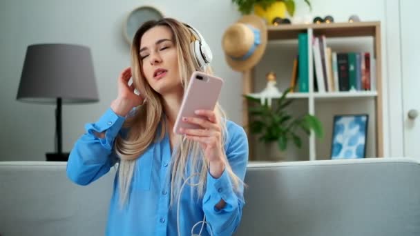 Portrait of young cute attractive young girl in cozy living room background listening to music with headphones. Woman wearing blue blouse enjoying the music and dancing at home. - Video