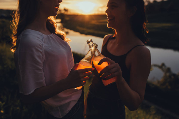 Two best friends holding a bottle, Girls having fun with a cocktail at sunset, positive face expression, outdoor, concept of joy - Photo, image