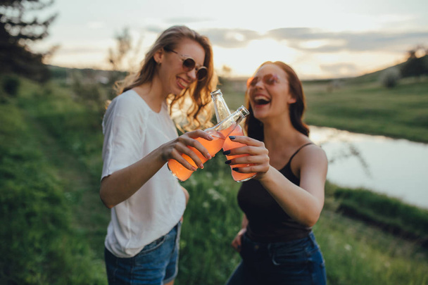 deux amies heureuses, tenant une bouteille dans sa main, dans des lunettes de soleil, se concentrer sur une bouteille, au coucher du soleil, expression faciale positive, concept extérieur, vacances et bonheur, Defocus
 - Photo, image