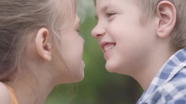 Close up portrait of cute boy and pretty girl sitting in the park, trying to rub their noses and having fun. A couple of happy children. Funny carefree kids in love outdoors. - Materiał filmowy, wideo