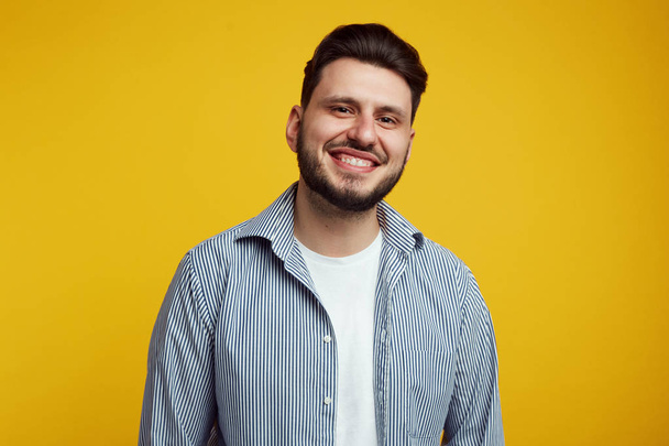 Çekici sakallı genç adam yüksek sesle gülüyor, geniş gülümseyerek, sarı arka plan karşı onun beyaz düz dişleri gösteren - Fotoğraf, Görsel
