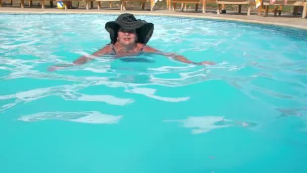 Şapkalı beyaz yaşlı kadın yaşlı bir otelde mavi su havuzunda yüzer. Sağlıklı bir yaşam tarzı kavramı, emeklilik te aktivite. Yavaş Çekim - Video, Çekim