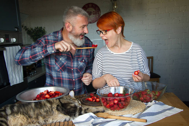 Para dorosłych mężczyzna i kobieta skórki i cięte truskawki na dżem truskawkowy, karmić się nawzajem, śmiech i baw się, kociaka Maine Coon jest na stole w kuchni - Zdjęcie, obraz