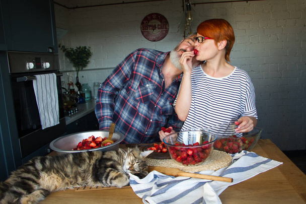 Para dorosłych mężczyzna i kobieta skórki i cięte truskawki na dżem truskawkowy, karmić się nawzajem, śmiech i baw się, kociaka Maine Coon jest na stole w kuchni - Zdjęcie, obraz