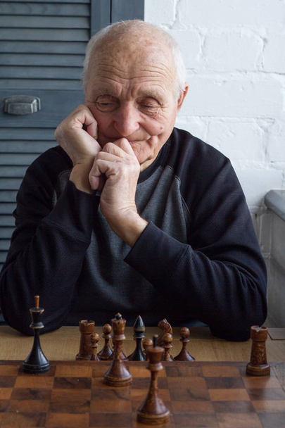 Um homem velho está sentado na frente de um tabuleiro de xadrez, pensando sobre o próximo movimento em um jogo de xadrez
 - Foto, Imagem
