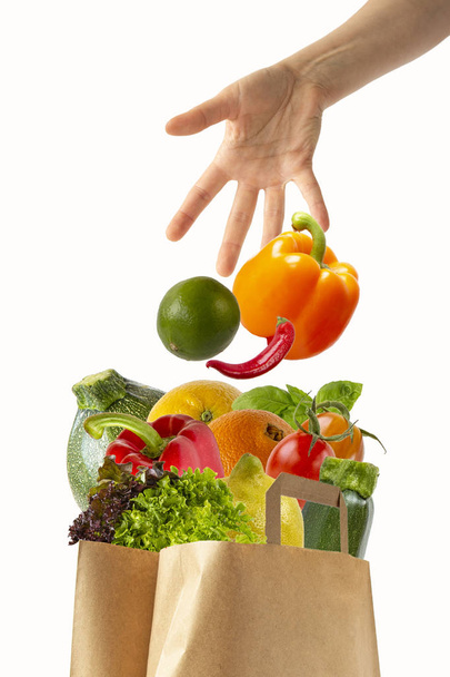 Female hand throwing vegetables in a paper bag with food.Isolated objects on white background. - Photo, Image
