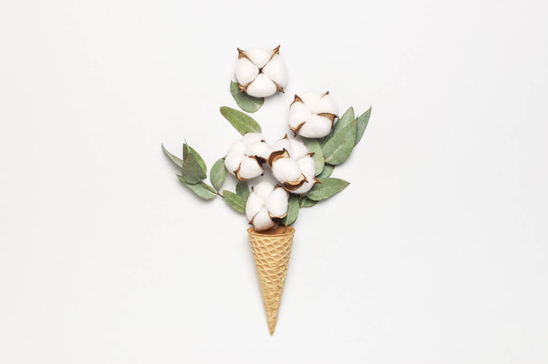 Waffle cone with bouquet of cotton flowers and fresh eucalyptus twigs on light gray background. Creative Delicate white cotton flowers minimal concept. Flat lay top view copy space. - Fotó, kép