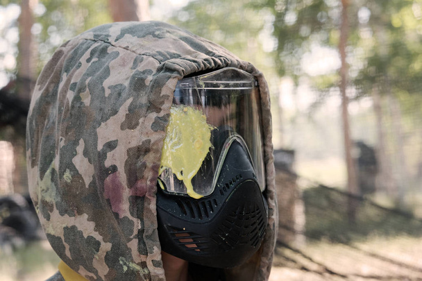 Paintball oyuncusunun kafa atışı. Profildeki adam, ormandaki camın üzerinde sarı bir leke olan koruyucu maskeli kamuflaj başlığıyla. Spor yaşam tarzı veya eğlence ve takım savaş oyunları. - Fotoğraf, Görsel