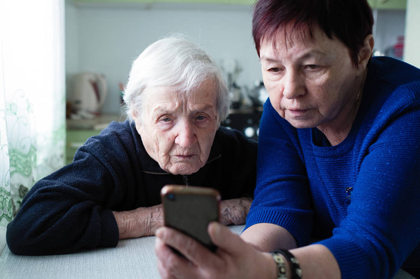 Μια ενήλικη γυναίκα δείχνει μια γριά γυναίκα την οθόνη του smartphone. - Φωτογραφία, εικόνα