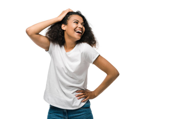 feliz rizado africano americano mujer tocando el pelo, mientras que de pie con la mano en la cadera aislado en blanco
  - Foto, Imagen