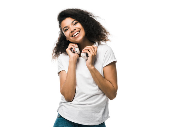 boldog afro-amerikai lány tartja a fejhallgatót, miközben nézi kamera izolált fehér  - Fotó, kép