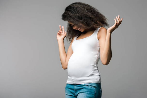cheerful pregnant african american girl looking at belly on grey  - Фото, зображення