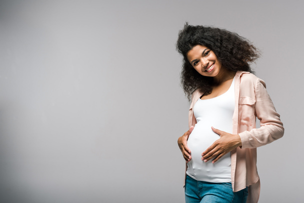 positive young pregnant african american woman touching belly on grey  - 写真・画像