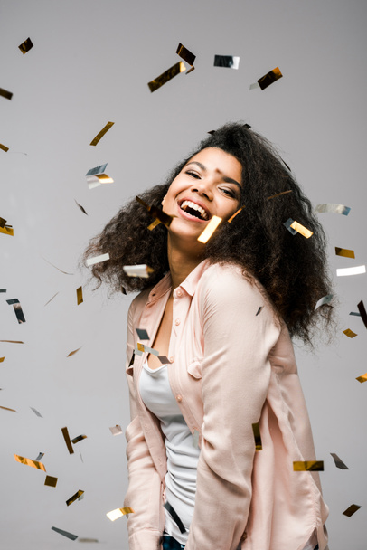 cheerful african american girl smiling near shiny confetti on grey  - Photo, Image