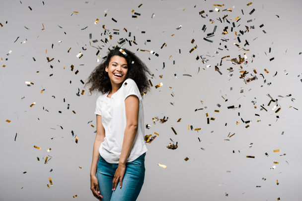 heureuse jeune femme afro-américaine souriant près de confettis brillants tout en se tenant sur gris
  - Photo, image