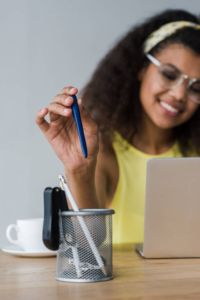 selective focus of pen holder and happy african american woman taking pen while using laptop   - Photo, Image