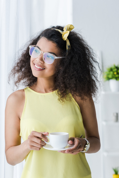 cheerful african american woman in glasses holding saucer and cup with drink  - Foto, Imagem
