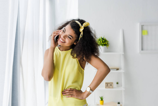 cheerful african american woman talking on smartphone while standing with hand on hip  - Foto, Imagem