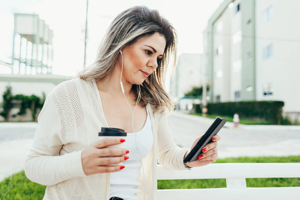 Nuori nainen istuu penkillä, pitää kahvikuppia kädessään ja lukee e-kirjaa kaupungissa
 - Valokuva, kuva