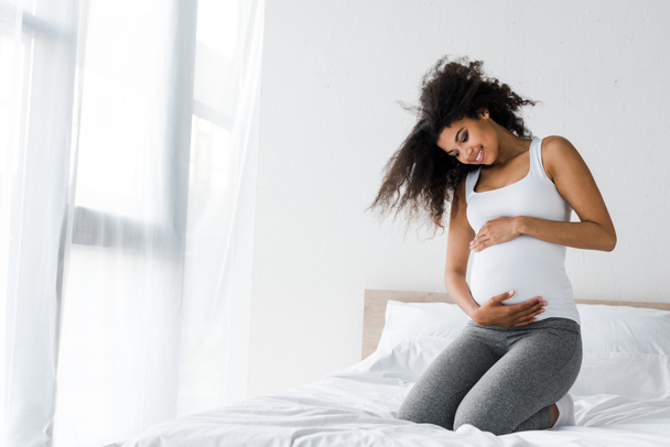 heureux enceinte africaine américaine toucher le ventre tout en étant assis sur le lit
  - Photo, image