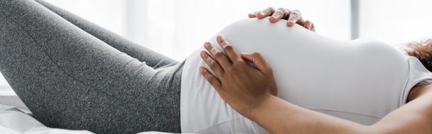 Panoramaaufnahme einer schwangeren Afroamerikanerin, die ihren Bauch berührt, während sie auf dem Bett liegt  - Foto, Bild