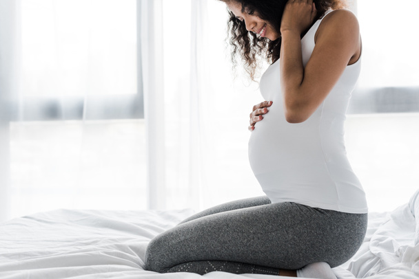 recortado vista de feliz rizado embarazada africana americana tocando vientre mientras está sentado en la cama
  - Foto, imagen