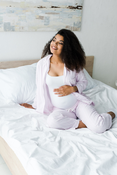 positive young pregnant african american woman sitting on bed and touching belly  - Valokuva, kuva