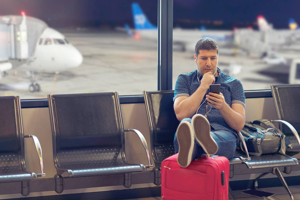 Średnim wieku człowiek turysta za pomocą inteligentnego telefonu w terminalu lotniska patrząc na harmonogramy lotów, podróżny siedzi w międzynarodowym terminalu wylotu za pomocą telefonu, aby sprawdzić anulowane opóźnienia lotnicze - Zdjęcie, obraz