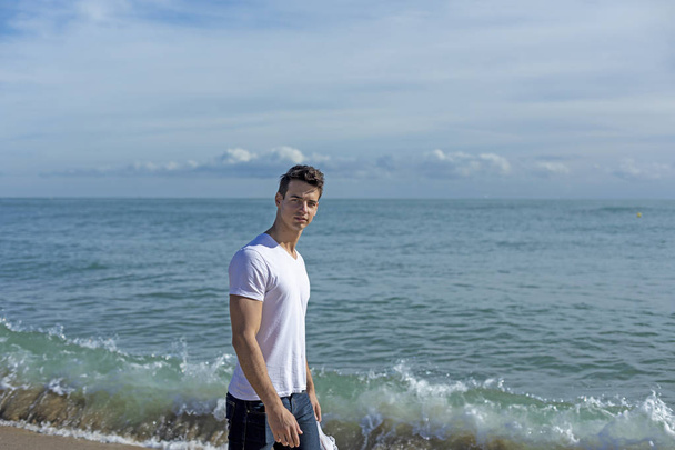 Νεαρός ελκυστικός άντρας περπατά στην παραλία κρατώντας ένα ζευγάρι αθλητικά παπούτσια σε μια ηλιόλουστη μέρα - Φωτογραφία, εικόνα