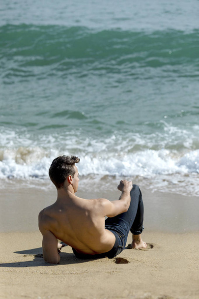 Вид сзади на молодого обнаженного мужчину в джинсах, сидящего на берегу моря и смотрящего в солнечный день
 - Фото, изображение