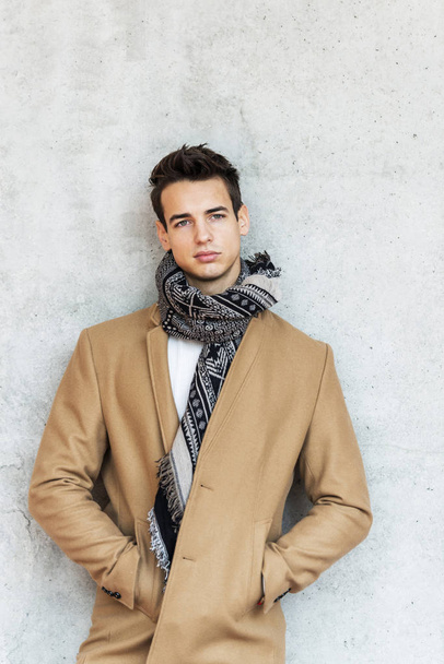 Front view of stylish young man wearing coat and scarf leaning on a wall while looking away - Foto, immagini