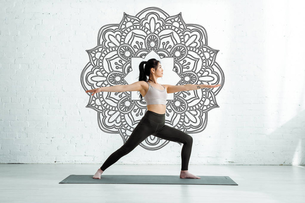 спокійна азіатська жінка стоїть з простягнутими руками на йога килимок біля мандали орнаменту
  - Фото, зображення