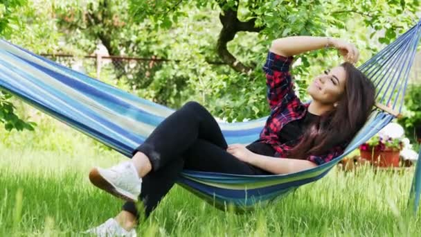 Hermosa joven feliz se encuentra en una hamaca en el jardín, sonríe y se relaja
 - Metraje, vídeo