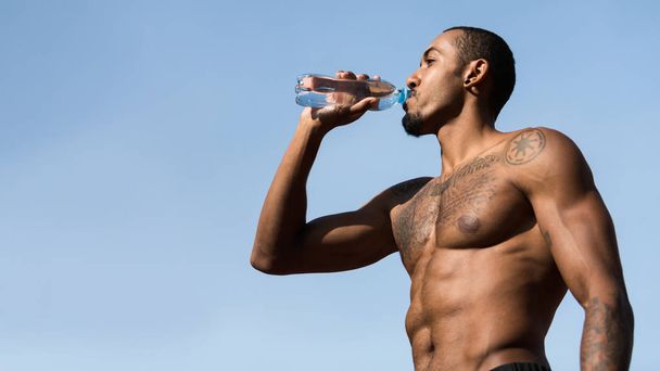 Мускулистый афро-человек пьет воду после тренировки против голубого неба
 - Фото, изображение