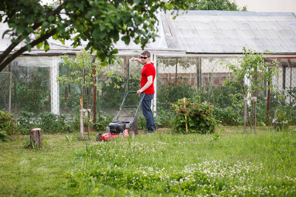 Young man in red t-shirt and sunglasses mows the grass with a grass-cutter on the lawn in the backyard in the village on a sunny summer day. - Fotó, kép