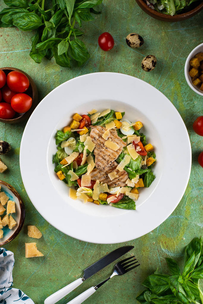 frischer Snack-Salat mit gegrillter Hühnerbrust und Parmesan auf weißem Teller auf grünem schäbigen Tisch mit Zutaten - Foto, Bild