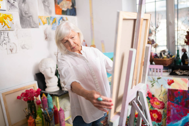 Aged artist with short blonde hair standing near drawing easel - Фото, зображення