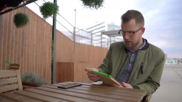 Młody mężczyzna siedzi w kawiarni na świeżym powietrzu i używa tabletu - Materiał filmowy, wideo