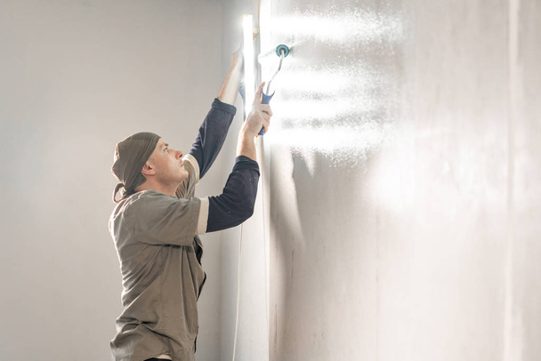 Young repairman smear on wall glue with a roller. . Worker glueing wallpapers on concrete wall. Repair the apartment. Home renovation concept. White Wallpaper for paint - Zdjęcie, obraz