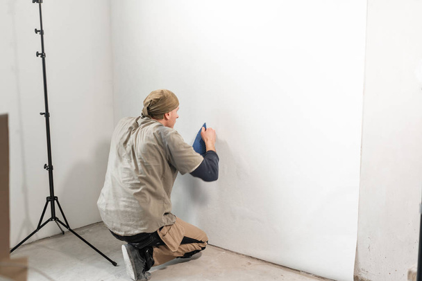 Young repairman aligns with plastic spatula. Worker glueing wallpapers on concrete wall. Repair the apartment. Home renovation concept. White Wallpaper for paint - Photo, Image