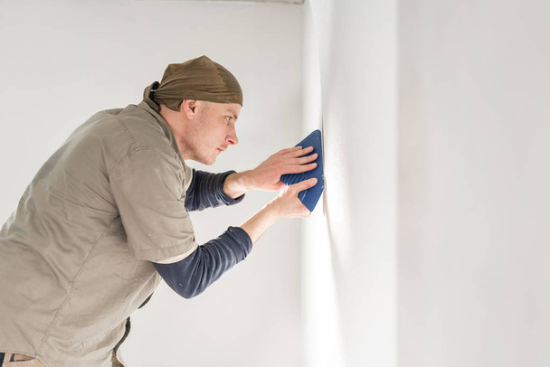 Young repairman aligns with plastic spatula. Worker glueing wallpapers on concrete wall. Repair the apartment. Home renovation concept. White Wallpaper for paint - Photo, image