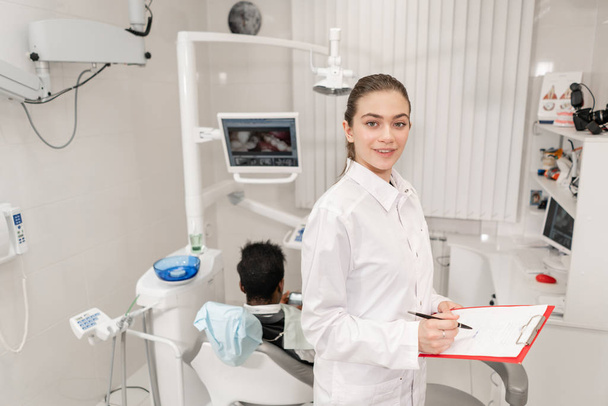 Charming beautiful girl dentist looks into the camera. Young African American male patient at chair at dental clinic. Medicine, health, stomatology concept. dentist conducts inspection and concludes. - Fotó, kép