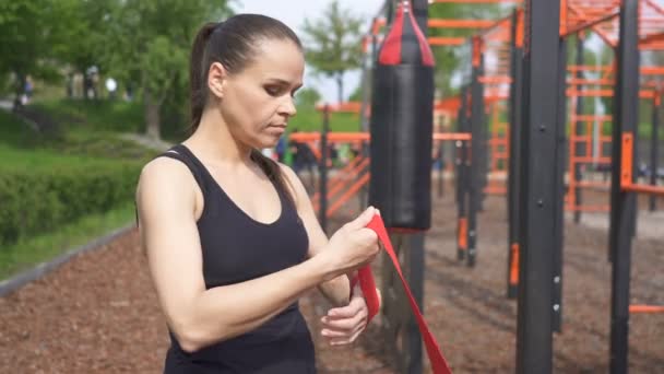 4k. ストリートトレーニンググラウンドでのトレーニングプロセスの準備をしている大人の女性ボクサー.手の赤いテープに巻き上げる - 映像、動画