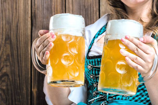 cropped shot of young woman in traditional bavarian costume holding beer glasses on wooden background - 写真・画像