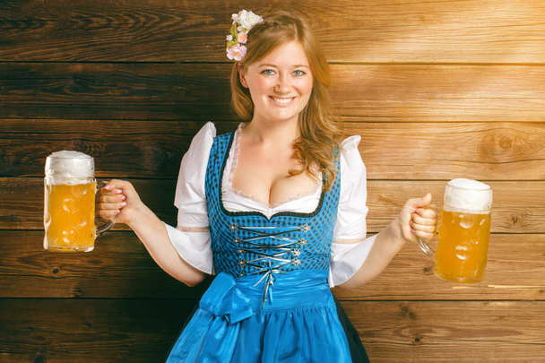 attractive young woman in traditional bavarian costume holding beer glasses and smiling at camera on wooden background  - Zdjęcie, obraz