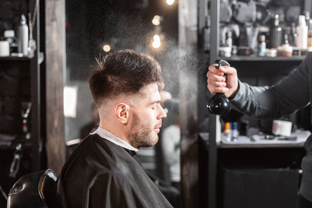 Barber sprays clean water on head in barbershop. Professional trimmer tool cuts beard and hair on young guy in barber shop salon. - Photo, Image