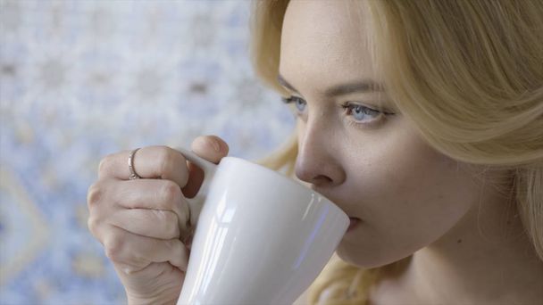 美しい青い目の若いブロンドの女性は、お茶を飲んでいます。アクション。白いマグカップから香りの良い熱いお茶を飲んで彼女の指に指輪を持つ若い美しい女性 - 写真・画像
